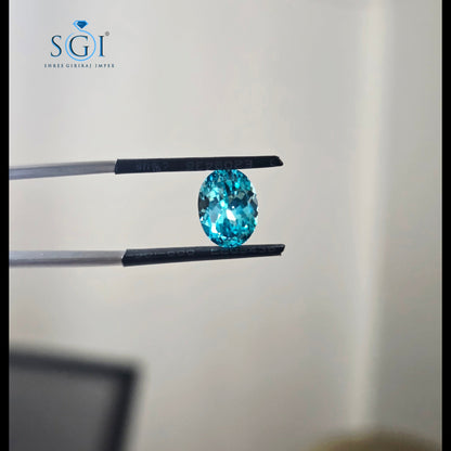 A round brilliant-cut light blue gemstone held in a pair of metal tweezers against a soft, out-of-focus light gray background. The SGI (Sapphire Global Impex) logo is visible in the upper left corner.